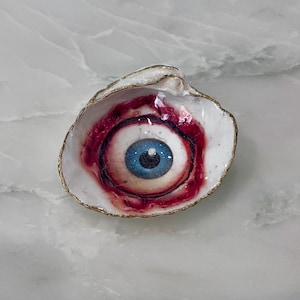 May include: A white seashell dish featuring a hand-painted realistic human eye with a bright blue iris and a black pupil. The eye is surrounded by deep red and pink bloodshot details. The outer edge of the shell is trimmed with a metallic gold finish.