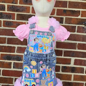 May include: A pink outfit for a child, featuring a pink top and denim overalls. The overalls are decorated with colorful cartoon characters, numbers, and a rainbow. The outfit is trimmed with pink tulle and includes a matching pink headband and sunglasses.