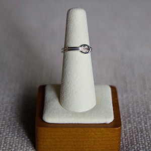 May include: A silver ring with a simple knot design, displayed on a white ring stand with a wooden base.