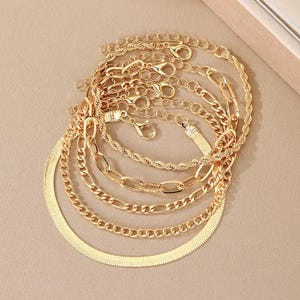 May include: A collection of gold-colored necklaces and bracelets. The jewelry features various chain styles, including herringbone, rope, and curb chains. Each piece has a clasp for easy wear. The pieces are arranged on a neutral surface.