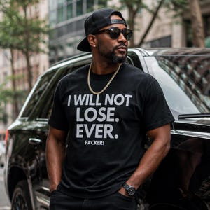 May include: A black short-sleeved t-shirt featuring the white text I WILL NOT LOSE. EVER. F#CKER! printed on the chest. The shirt is paired with a black baseball cap, dark sunglasses, a gold chain necklace, and a black wristwatch on the left wrist.