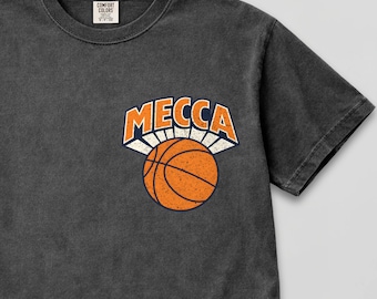 May include: Dark gray t-shirt with a crew neck and short sleeves. The shirt features a basketball design with the word "MECCA" in orange and white above an orange basketball. The shirt has a vintage, worn look.