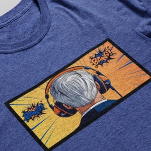 May include: Blue t-shirt featuring a pop art style graphic. The graphic shows a person wearing headphones, with the back of their head visible. The graphic includes the word "BANG!" in a comic book style.