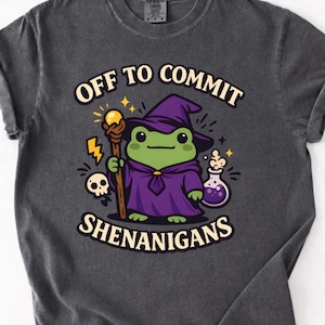 May include: Dark grey t-shirt with a cartoon frog wizard illustration. The frog wears a purple cloak and hat, holding a staff. Text reads "OFF TO COMMIT SHENANIGANS" above and below the frog design.