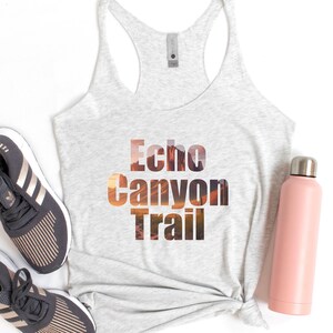 May include: A gray tank top with the text "Echo Canyon Trail" printed on the front. The text is layered with a sunset landscape image. The tank top is laying on a white surface with a pair of gray running shoes and a pink water bottle.