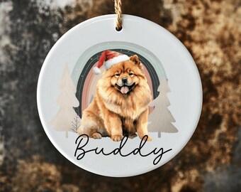 May include: White ceramic Christmas ornament featuring a brown and white Chow Chow dog wearing a red Santa hat. The dog is sitting in front of a rainbow and two stylized trees. The ornament is personalized with the name "Buddy".