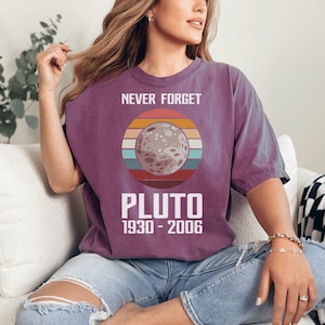 Könnte beinhalten: Ein lila T-Shirt mit einer Grafik von Pluto, mit einem Retro-Sonnenuntergangsdesign und dem Text "NEVER FORGET PLUTO 1930-2006". Das Shirt ist ein bequemer, lässiger Stil.
