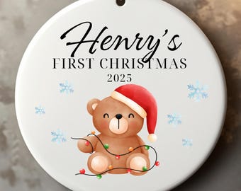 May include: A white Christmas ornament with the text "Henry's FIRST CHRISTMAS 2025". It features a brown teddy bear wearing a Santa hat and holding string lights. Blue snowflakes are scattered around the bear.