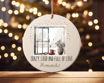 May include: White ceramic Christmas ornament with a window scene of a snowy house and a potted plant. The ornament has the text "CRAZY. LOUD AND FULL OF LOVE" and the name "Hernandez" written below.