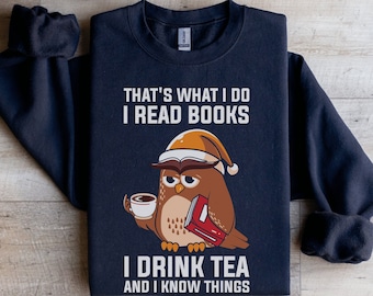 May include: Navy blue sweatshirt with a graphic of an owl wearing a Santa hat, holding a book and a cup of tea. The text on the sweatshirt reads "That's what I do I read books I drink tea and I know things."