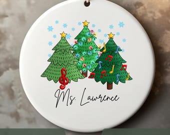 May include: A white ceramic Christmas ornament with a string for hanging. The ornament features three green Christmas trees with musical notes and snowflakes. The text "Ms. Lawrence" is written in black script.
