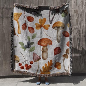 Può includere: Una coperta in tessuto intrecciato con sfondo bianco e un motivo autunnale colorato di funghi, foglie e bacche. La coperta ha frange su tutti i lati.