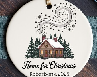 May include: A round, white Christmas ornament with a winter scene. A cozy cabin with a smoking chimney is surrounded by pine trees and snowflakes. The text "Home for Christmas" and "Robertsons, 2025" are displayed in elegant script.