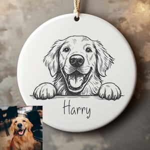 May include: White ceramic ornament with a black and white illustration of a Golden Retriever dog. The dog is smiling with its tongue out, and the name "Harry" is written below. A brown string is attached for hanging.