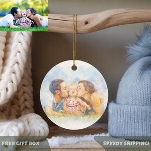 May include: A round ceramic ornament featuring a watercolor painting of a family kissing a baby. The ornament hangs from a gold string. The background includes a knitted scarf and a blue hat.