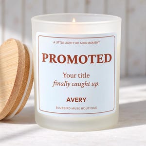May include: A frosted glass candle with a bamboo lid. The label features the text: A LITTLE LIGHT FOR A BIG MOMENT, PROMOTED, Your title finally caught up., AVERY, BLUEBIRD MUSE BOUTIQUE. The candle has a lit wick inside the glass container.