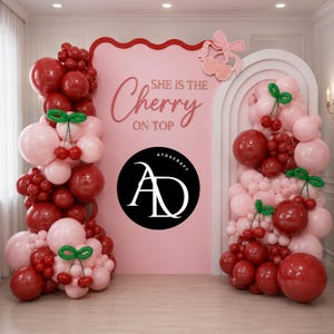Op de afbeelding: Een roze achtergrond met de tekst "She is the Cherry on Top" en een kersendesign. De achtergrond is versierd met rode en roze ballonnen, waarvan sommige de vorm van kersen hebben. Een zwarte cirkel met witte tekst en een logo staat in het midden.