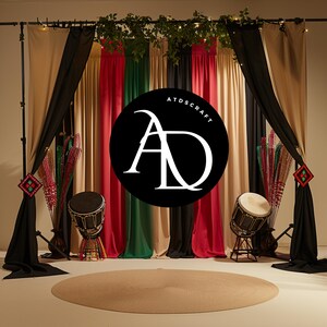 May include: A black and white logo with the letters "AD" in a circle, set against a backdrop of colorful fabric curtains. The curtains are in shades of beige, black, red, green, and beige. Two drums are placed in front of the curtains.