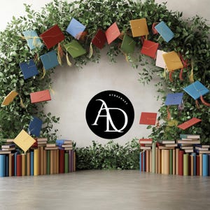 Puede incluir: Un logotipo en blanco y negro con las letras "AD" dentro de un círculo, con el texto "ATDSCRAFT" sobre las letras. El logotipo está rodeado de una guirnalda de gorros de graduación coloridos y follaje verde. Dos pilas de libros están a cada lado del logotipo.