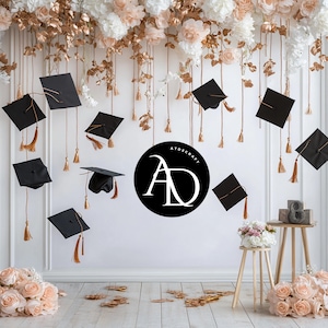 Może przedstawiać: Biała ściana ozdobiona czarnymi czapkami absolwentów wiszącymi na złotych frędzlach. Na ścianie znajduje się również czarne koło z białymi literami "AD" i tekstem "ATDSCRAFT" nad literami. Na małym drewnianym stoliku stoi biały wazon z kwiatami i drewniana cyfra "8".