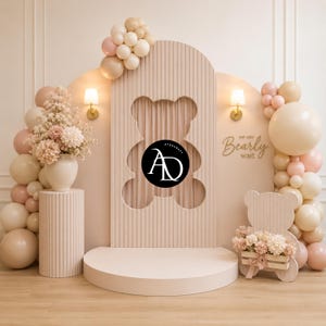 May include: A light pink and beige baby shower backdrop with a large teddy bear cutout, balloons, flowers, and the text "We can bearly wait." The backdrop includes a stage and decorative lighting, creating a festive atmosphere for a baby shower or similar event.