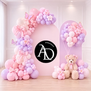 May include: A decorative balloon arrangement in pastel shades. The display includes a balloon arch with pink, lilac, and white balloons, bows, and floral details. A teddy bear with a pink bow sits near a matching backdrop, surrounded by balloons.