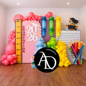 Könnte beinhalten: Eine Abschlussdekoration mit rosa Hintergrund, dem Jahr 2026, und einem Lineal. Es umfasst blaue, grüne und gelbe Buntstiftdekorationen, Ballongruppen, einen Bleistiftbecher und eine Eule mit Abschlusskappe. Auf einem Holzboden.