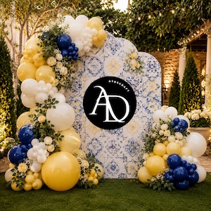 Op de afbeelding: Een decoratieve evenementenachtergrond met een blauw en geel tegelpatroon en een geschulpte bovenrand. Het display is versierd met een grote ballonslinger in wit, geel en marineblauw, geaccentueerd met witte rozen en groen blad.