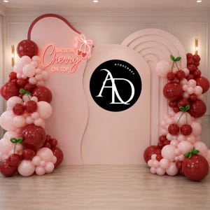 May include: A pink backdrop with cherry-themed decorations. The backdrop features a neon sign that reads "She is the Cherry on Top." Clusters of red and pink balloons are arranged to resemble cherries. A black circle with white text is also present.