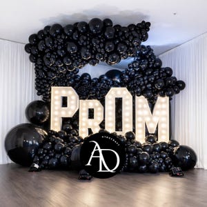 Puede incluir: Una exhibición decorativa para un evento de graduación. La palabra "PROM" está escrita en letras grandes e iluminadas. Las letras están rodeadas por una gran disposición de globos negros. Un logotipo es visible en la parte inferior.