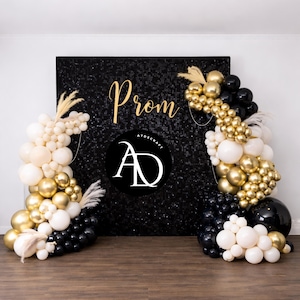 Peut inclure: Un fond noir à paillettes avec le mot "Prom" en lettres dorées, flanqué d'arches de ballons noirs, dorés et crème. Les arches sont décorées d'éléments floraux séchés. Un cercle noir avec du texte blanc est centré sur le fond.
