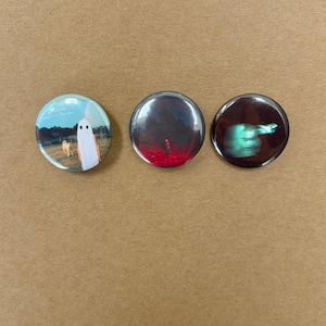 May include: Three circular pin-back badges featuring distinct photographic prints. The first shows a ghost figure and a dog in a field. The second displays a dark landscape with red ground. The third depicts a blurred green light on a dark background.