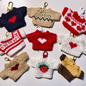 Puede incluir: Diez llaveros de punto con diferentes patrones y colores. Los suéteres son todos pequeños y tienen un diseño de corazón o fresa. Los llaveros están hechos de hilo y tienen un anillo de metal dorado.