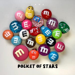 Può includere: Una collezione di ciondoli rotondi e colorati a forma di caramella con la lettera "m" stampata su di essi. I ciondoli sono disposti in cerchio su una superficie bianca. Il testo "JANE" e "POCKET OF STARS" è stampato sulla superficie.