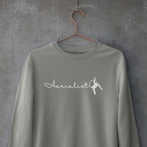 May include: A grey sweatshirt with the word "Aerialist" written in white cursive script and a white silhouette of a person on an aerial hoop. The sweatshirt is hanging on a hanger against a grey textured wall. The text "Graphite Heather" is written below the sweatshirt.