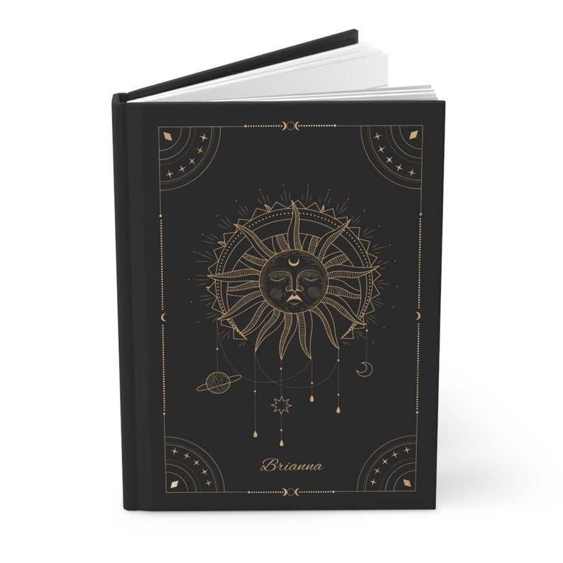May include: Black hardcover journal with a gold sun and moon design. The journal has a gold border and the name "Brianna" is printed in gold on the front cover.