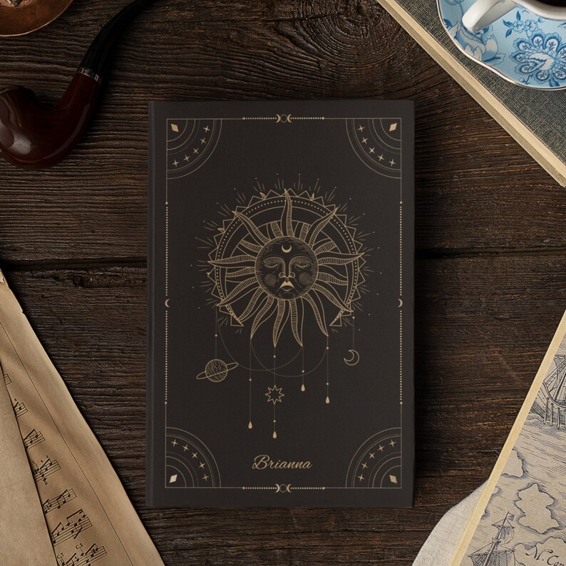 May include: Black journal with a gold sun and moon design. The journal has the name "Brianna" printed on the front.