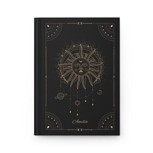 May include: Black journal with a gold sun and celestial design. The sun has a face and is surrounded by a circle of rays. The journal has the name "Amelia" written on the front.