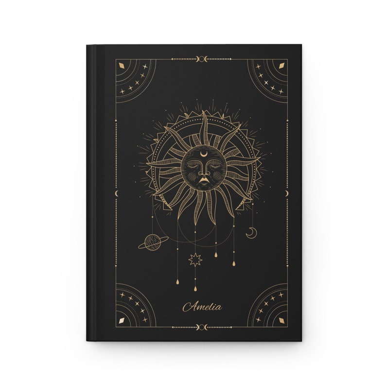 May include: Black journal with a gold sun and celestial design. The sun has a face and is surrounded by a circle of rays. The journal has the name "Amelia" written on the front.