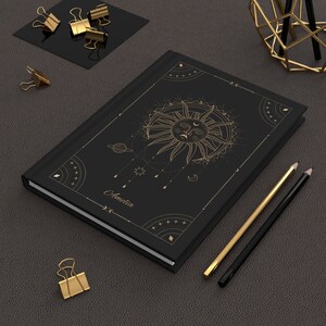 May include: Black notebook with a gold sun and moon design. The notebook has the name "Amelia" written on the front.