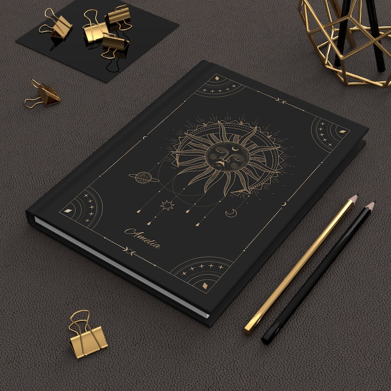 May include: Black notebook with a gold sun and moon design. The notebook has the name "Amelia" written on the front.
