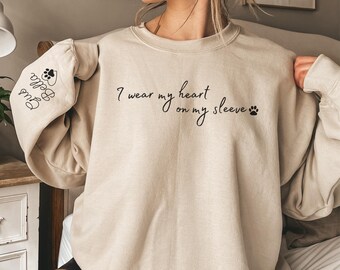 May include: A beige sweatshirt with the text "I wear my heart on my sleeve" and a paw print on the right side. The left sleeve has the text "Paw Prints" and a paw print.