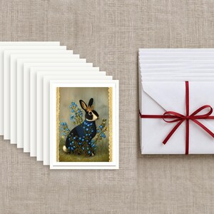 May include: A set of greeting cards featuring a black rabbit wearing a gold crown, surrounded by blue flowers, alongside a stack of white envelopes tied with a thin red ribbon. The cards have a gold border and a white frame.
