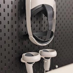 Op de afbeelding: Een witte virtual reality headset en twee witte controllers hangen aan een zwarte pegboard muur.