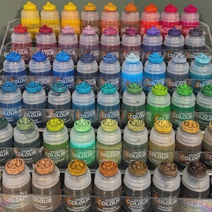 May include: A collection of small paint bottles arranged on a tiered shelf. The bottles are all the same size and shape, and they are filled with different colours of paint. The bottles are labelled with the words "Citadel Colour" and the name of the colour.