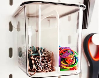 May include: A clear plastic container with a divider, holding copper paper clips and colorful rubber bands. The container is mounted on a white pegboard.