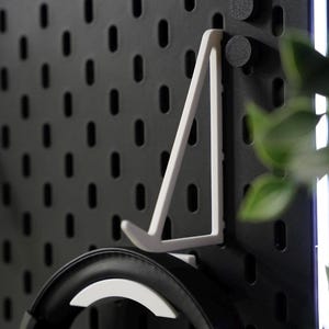 Puede incluir: Un tablero de clavijas negro con un soporte para auriculares blanco y unos auriculares negros colgados. El tablero de clavijas tiene una tira de luz blanca a lo largo del borde superior.