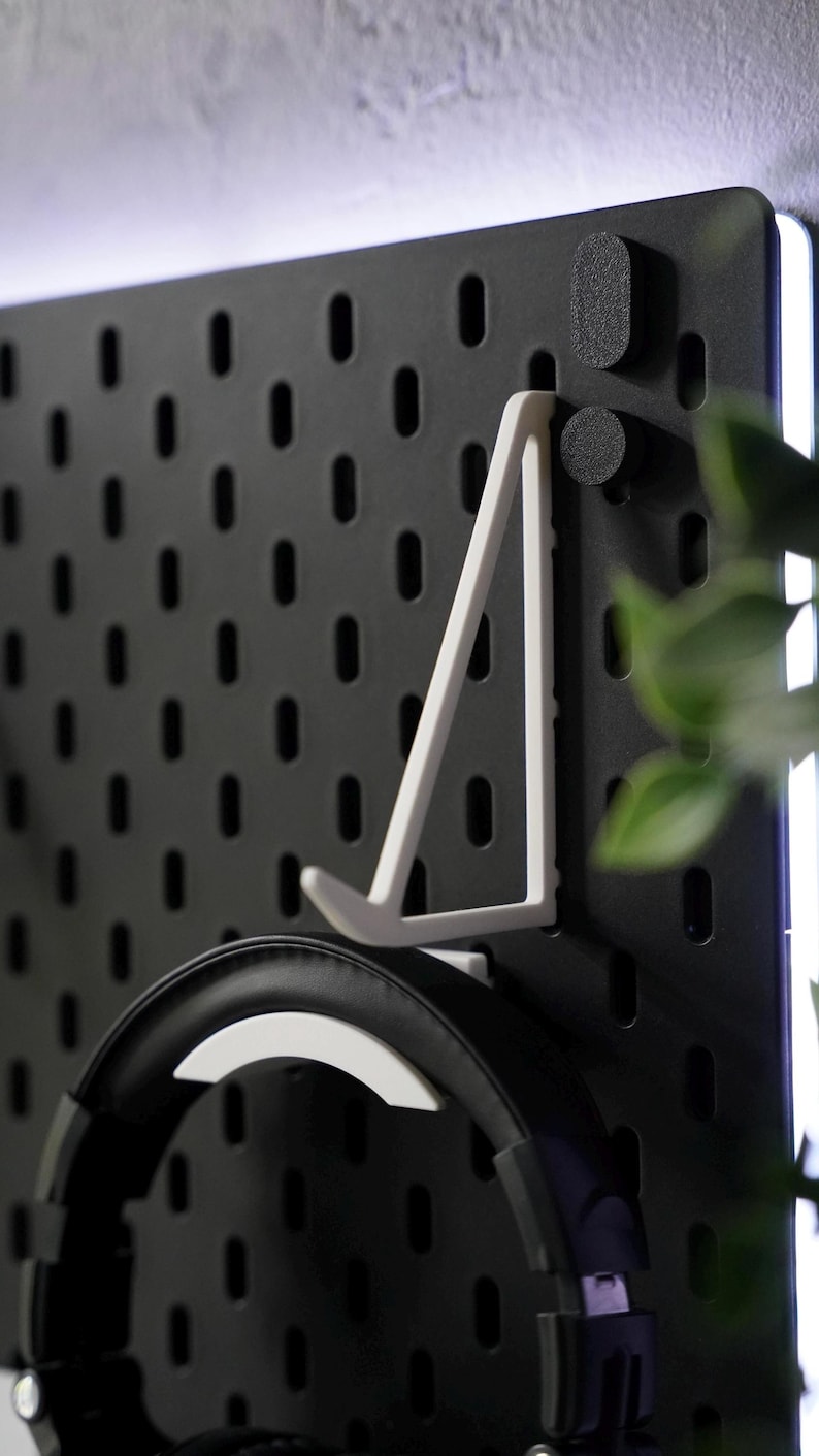 Puede incluir: Un tablero de clavijas negro con un soporte para auriculares blanco y unos auriculares negros colgados. El tablero de clavijas tiene una tira de luz blanca a lo largo del borde superior.