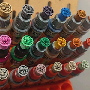 Op de afbeelding: Een verzameling kleine flesjes verf met kleurrijke, getextureerde dopjes. De dopjes zijn gevormd als dobbelstenen met verschillende kleuren en ontwerpen.