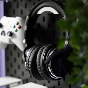 Puede incluir: Auriculares negros de diadema con diadema plateada colgados en una pared de tablero perforado. Tambi&eacute;n se puede ver un mando de videojuegos blanco y dos recipientes de pl&aacute;stico transparente con tapas blancas.